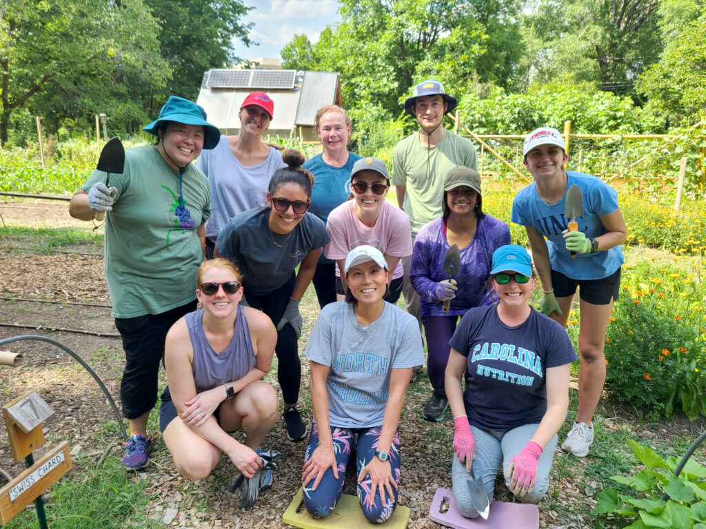 Group of smiling people posing together in a garden; several people are holding trowels and wearing gardening gloves